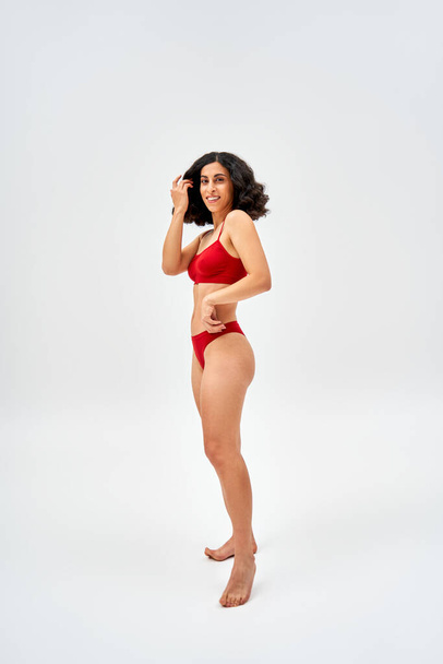 Full length of barefoot and middle eastern woman in modern red lingerie touching hair and looking at camera while standing on grey background, self-acceptance and body positive concept  - Foto, immagini