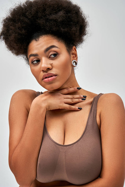 Portrait of confident and sexy african american woman in brown bra touching neck and looking away while standing and posing isolated on grey, self-acceptance and body positive concept  - Valokuva, kuva