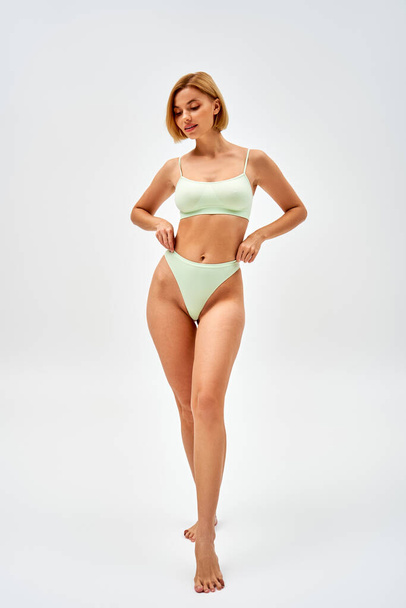 Full length of barefoot and blonde young woman in light green lingerie touching panties while standing and posing on grey background, self-acceptance and body positive concept  - Φωτογραφία, εικόνα