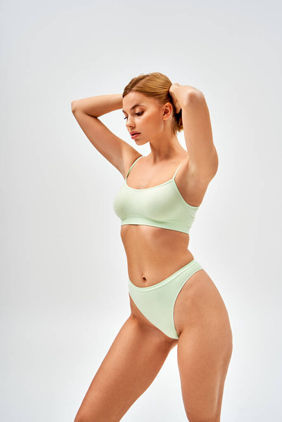 Sensual young woman with natural makeup wearing light green lingerie and touching blonde hair while looking down and standing isolated on grey, self-acceptance and body positive concept  - Photo, Image