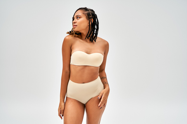 Modern young african american woman with natural makeup wearing beige bra and panties while touching hip isolated on grey, self-acceptance and body positive concept, femininity  - Foto, immagini