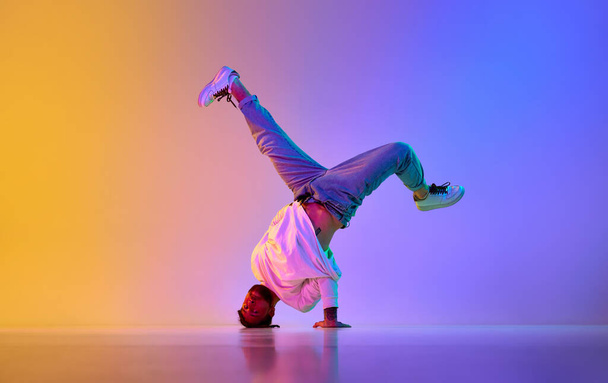 Creative young guy in casual clothes dancing hip-hop contemporary, breakdance against gradient studio background in neon light. Concept of street style dance, fashion, youth, hobby, dynamics, ad - Foto, Bild
