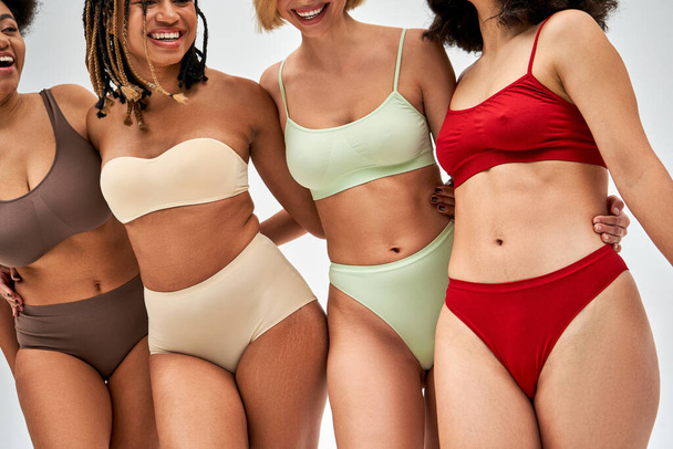 Cropped view of cheerful multiethnic women in colorful and modern lingerie standing and hugging each other isolated on grey, multicultural models and body positivity movement concept - Photo, Image