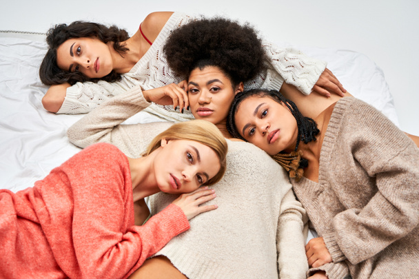 Высокий угол обзора модных и многоэтнических женщин в теплых и вязаных свитерах, лежащих вместе на кровати изолированных на сером, различных типах тела и самовосприятия, мультикультурного представления - Фото, изображение