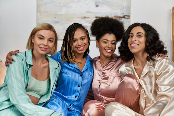 Portrait of positive african american women hugging multiethnic girlfriends in colorful pajama and looking at camera during pajama party, bonding time in comfortable sleepwear, slumber party - 写真・画像