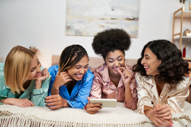 Laughing and multiethnic girlfriends in colorful pajama having fun while using smartphone together while lying on bed during pajama party at home, bonding time in comfortable sleepwear - Fotografie, Obrázek