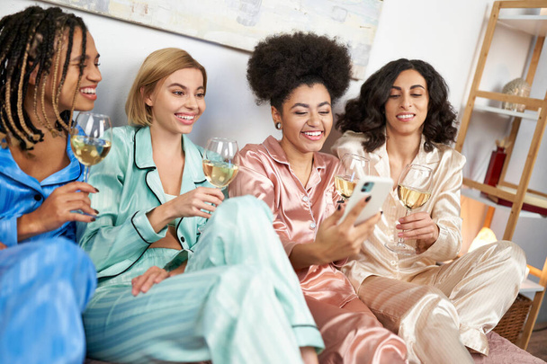 Positive and multiethnic girlfriends in colorful pajama using smartphone and holding glasses of wine while sitting on bed during girls night at home, comfortable sleepwear, slumber party - Fotografie, Obrázek