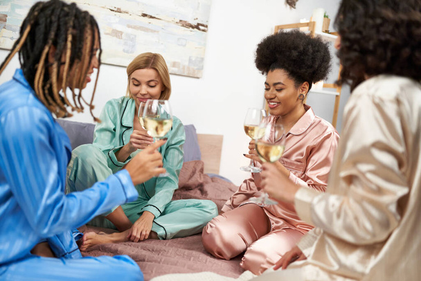 Smiling african american woman holding glass of wine while talking to multiethnic girlfriends with glasses of wine during girls night in bedroom at home, bonding time in comfortable sleepwear - Foto, imagen