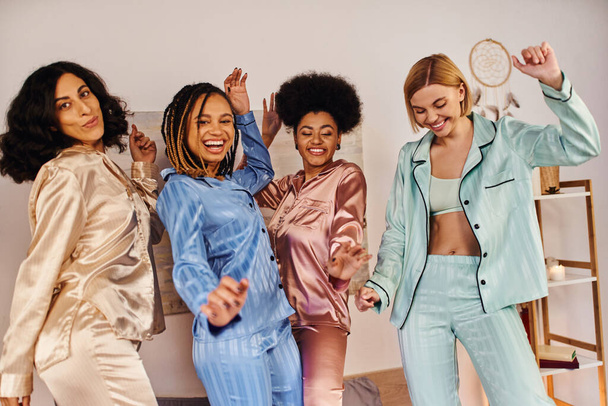Cheerful multicultural girlfriends in colorful pajama dancing and having fun together while looking at camera during pajama party at home, slumber party, bonding time in comfortable sleepwear - 写真・画像