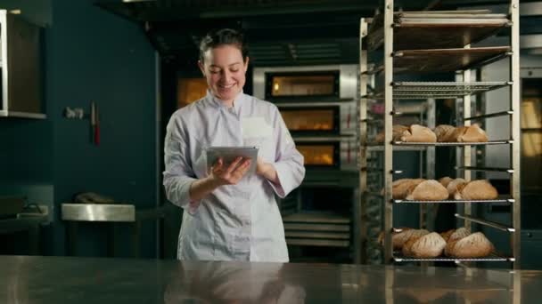 a woman baker in a uniform near the shelves with freshly baked bread holds tablet in her hands checks bread bakery - Footage, Video