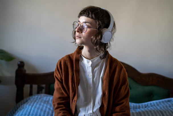 Sad pensive teenage girl wearing headphones sitting on bed looking aside, upset millennial woman listening music at home to boost mood and alleviate sadness. Podcasting in education - Fotoğraf, Görsel