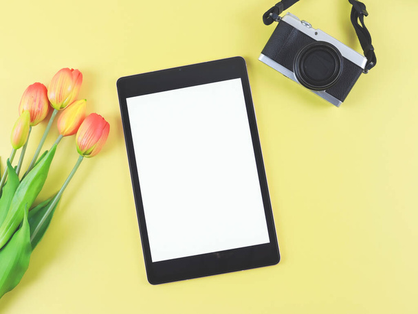 Top view vagy lapos laikus digitális tabletta üres fehér képernyő, tulipán virágok és digitális fényképezőgép izolált sárga alapon. - Fotó, kép