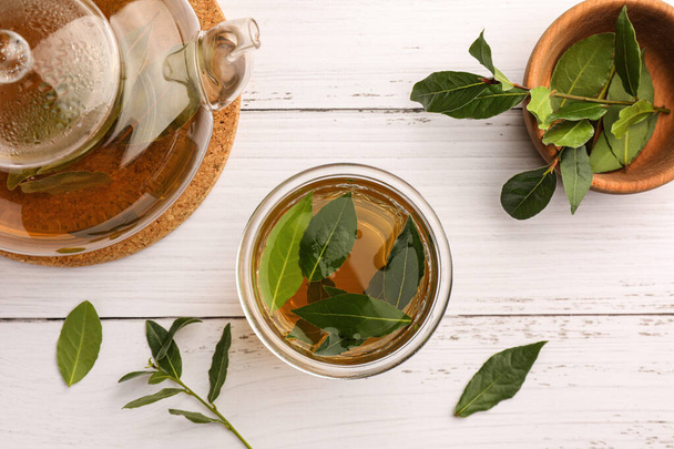 Cup of freshly brewed tea, teapot and bay leaves on white wooden table, flat lay - Photo, image