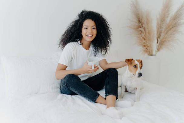 La joven feliz con peinado afro se relaja con su perro en una cama, disfrutando de bebidas calientes y expresando emociones positivas. Concepto de personas y descanso. - Foto, imagen