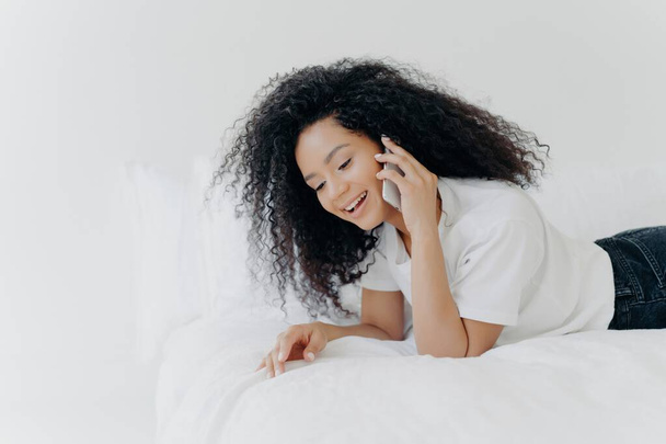 Relaxed woman in bed chats on phone, friendly convo before sleep, wearing white tee, cozy vibes. - Фото, изображение