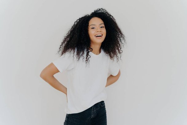 Femme afro-américaine joyeuse en vêtements décontractés, riant les mains derrière le dos, isolée sur fond blanc. - Photo, image
