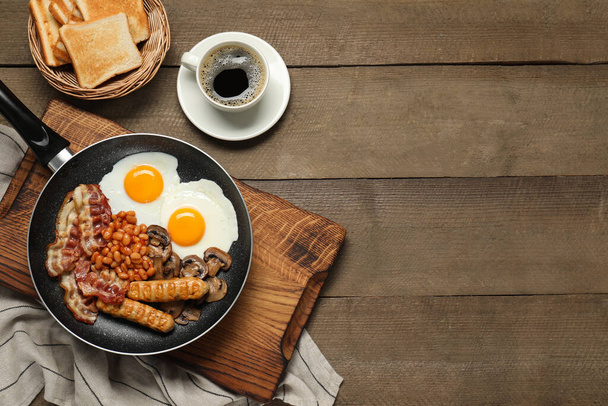 Frying pan with cooked traditional English breakfast and cup of coffee on wooden table, flat lay. Space for text - Foto, Imagen