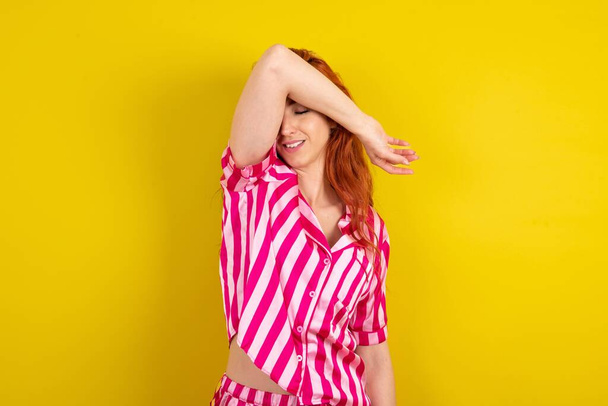 Jonge roodharige vrouw met een roze pyjama over een gele studioachtergrond die de ogen bedekt met een lachende arm, vrolijk en grappig. Blind concept. - Foto, afbeelding