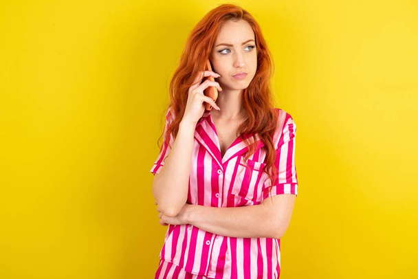 若い赤毛の悲しい女性は,スマートフォンで話す黄色いスタジオの背景の上にピンクのパジャマを着ています. コミュニケーションコンセプト. - 写真・画像