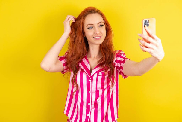 黄色いスタジオの背景にピンクのピンクのパジャマを着て,彼女のソーシャルメディアにそれを投稿する準備ができてセルフィーを取る若い赤毛の女性. - 写真・画像