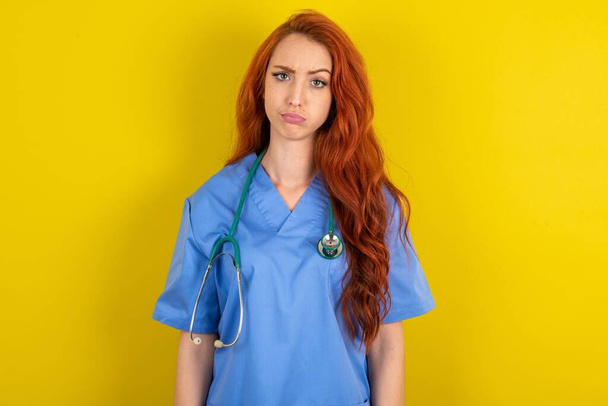 Nespokojený mladý rusovlasý lékař žena přes žluté pozadí studia mračí tvář cítí nešťastný má nějaké problémy. Koncept negativních emocí a pocitů - Fotografie, Obrázek