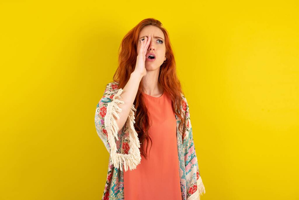 червона волохата жінка стоїть над жовтим студійним фоном, кричить і кричить голосно на бік з рукою на рот. Концепція комунікації. - Фото, зображення