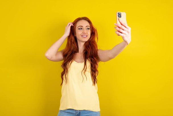 красива руда волохата жінка в жовтій сорочці над жовтим студійним фоном посміхається і приймає селфі, готовий розмістити його в своїх соціальних мережах. - Фото, зображення