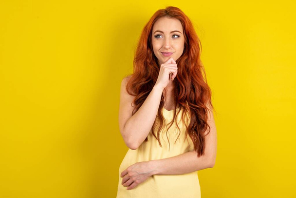 Thoughtful bela mulher de cabelos vermelhos vestindo camisa amarela sobre o fundo do estúdio amarelo mantém o queixo e olha para longe pensivamente faz grande plano - Foto, Imagem