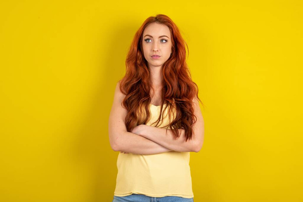 Charmante réfléchie belle femme aux cheveux rouges portant une chemise jaune sur fond de studio jaune se tient avec les bras pliés concentrés quelque part avec une expression coûteuse pense quoi faire - Photo, image