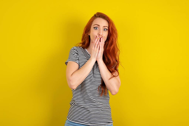 red haired woman wearing striped shirt over yellow studio background keeps hands on mouth, looks with eyes full of disbelief, being puzzled with amount of work - Foto, afbeelding