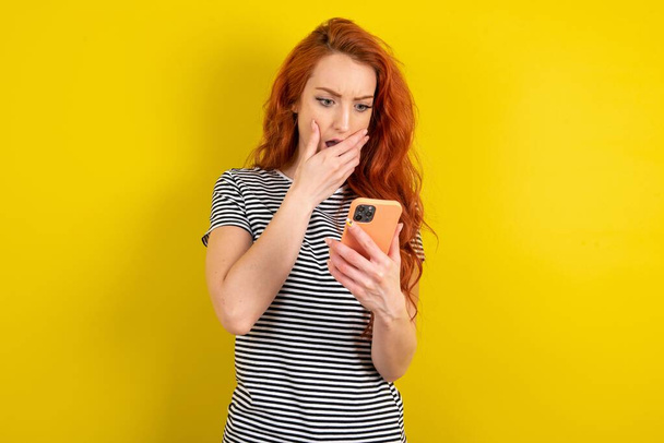 punatukkainen nainen yllään raidallinen paita yli keltainen studio tausta on syvästi yllättynyt, tuijottaa älypuhelimen näyttö, lukee järkyttäviä uutisia verkkosivuilla, Omg, sen kauhea! - Valokuva, kuva