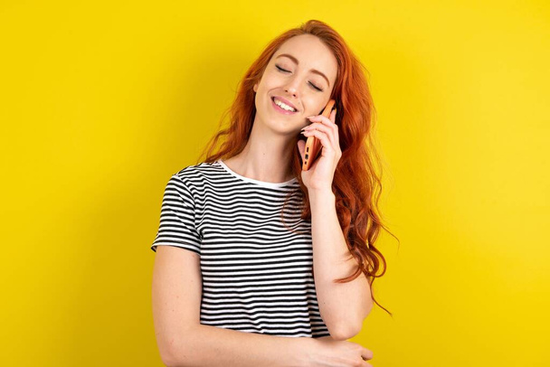 Funny red haired woman wearing striped shirt over yellow studio background laughs happily, has phone conversation, being amused by friend, closes eyes. - Fotografie, Obrázek