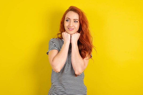 femme aux cheveux rouges portant une chemise rayée sur fond jaune studio tient la main sous le menton, heureux d'entendre des mots chaleureux de l'étranger - Photo, image