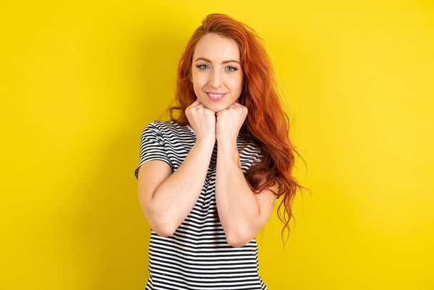 Sarı stüdyo arka planında çizgili tişört giyen kızıl saçlı kadın çeneye iki eliyle dokunur, güzel gülümser, sevgilisiyle mutlu bir gün geçirir. - Fotoğraf, Görsel