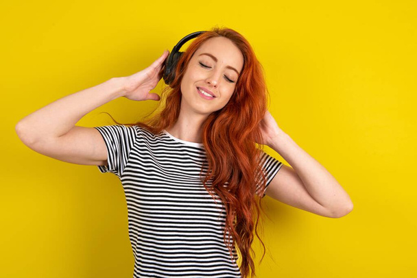 rothaarige Frau mit gestreiftem Hemd über gelbem Studiohintergrund lächelt breit und fühlt sich sehr froh, Lieblingsmusik über drahtlose Kopfhörer zu hören schließt die Augen. - Foto, Bild