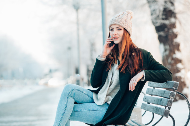 young woman in winter park talking mobile phone, sms, sitting on the bench - Foto, Bild