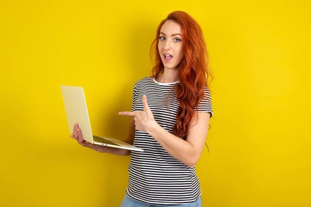 Shocked red haired woman wearing striped shirt over yellow studio background pointing finger modern device - Foto, Imagem