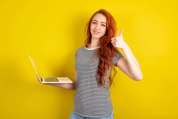 Bella attraente donna dai capelli rossi indossa camicia a righe su sfondo giallo studio tenere netbook mano dito mostrare pollice in su - Foto, immagini