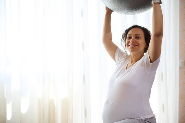 Affascinante donna incinta con grande pancia in 28 settimane di gravidanza, esercizio con un fitball a casa, sorridente guardando la fotocamera. Copia spazio pubblicitario. Fitness prenatale. Stile di vita sano concetto attivo - Foto, immagini