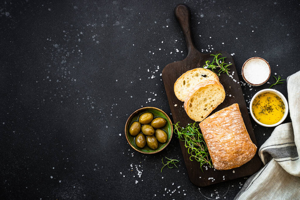 Pane ciabatta su tavola di legno con olio d'oliva, olive ed erbe aromatiche su nero. Vista dall'alto con spazio di copia. - Foto, immagini
