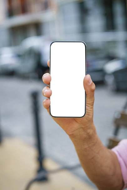 Close-up of mans hand holding mobile phone with white screen display, offering ample copy space. Background features urban cityscape, providing modern and vibrant backdrop. . High quality photo - Foto, Bild