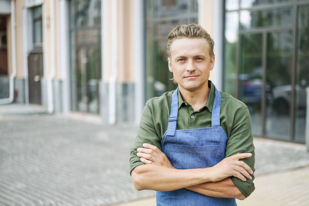 Restaurant or kitchen worker wearing blue apron, standing against backdrop of city. Concept of service and readiness to help. Customer service-oriented nature of the hospitality industry. . High - Foto, Imagen