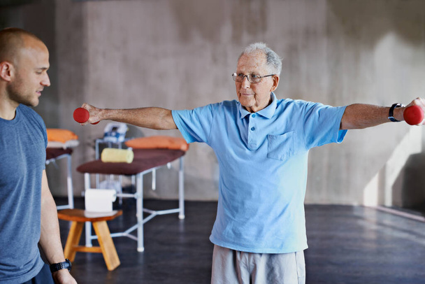 Fizioterapeuta segítség, idős férfi és súlyzós képzés az egészség- és wellness terápiára nyugdíjba vonuláskor. Egészségügyi ellátás, fizio és edzés a gyógyuláshoz súlyzóval az orvosi gyakorlat és az idősek számára. - Fotó, kép