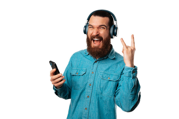 Homem barbudo animado está mostrando o gesto de rock enquanto ouve a música sobre fundo branco. - Foto, Imagem