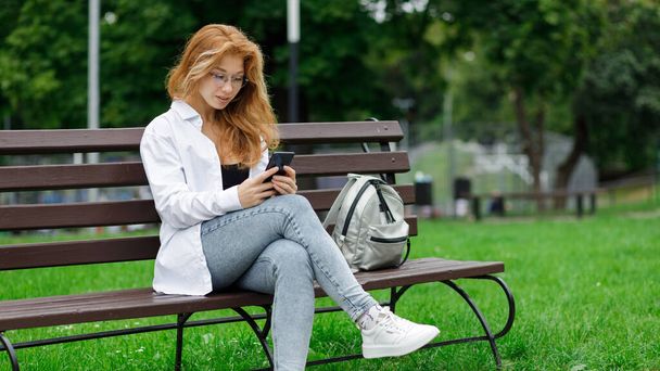 Young beautiful woman using her smartphone sitting on the bench in the park. - Foto, Bild