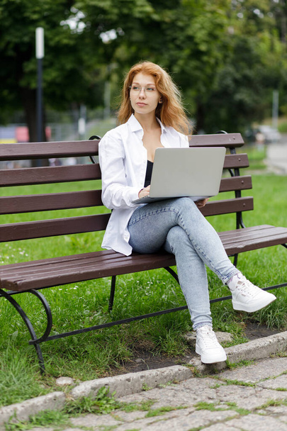 Young confident attractive student girl using her laptop sitting on the bench in park. - Foto, afbeelding