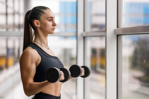 Young athletic fitness girl doing biceps curls with dumbbells near the window. - Foto, Bild
