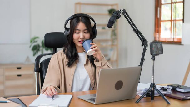 Kobieta podcaster picia kawy i pisania notatki na papierze podczas pracy do nagrywania podcastu wideo. - Zdjęcie, obraz