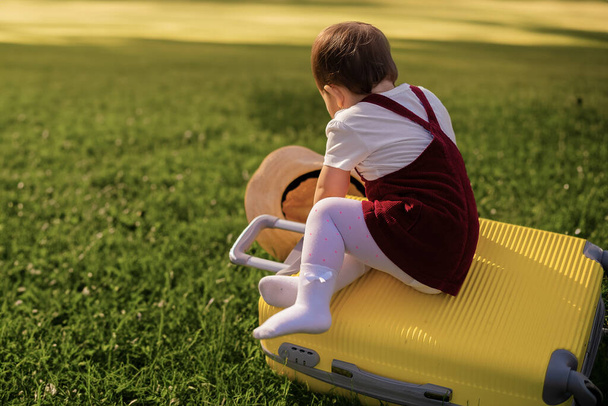 Ребенок сидит на чемодане и, закрыв глаза, мечтает о путешествии, приключениях, отпуске. Маленькая девочка на фоне зеленой травы - Фото, изображение