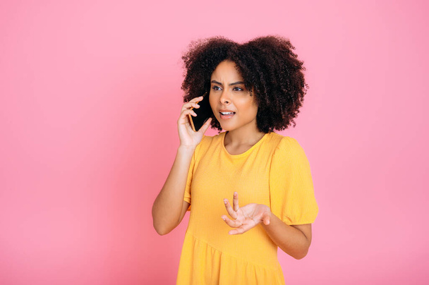 Phone talk. Confused successful busy mixed race curly haired woman, talking on mobile phone with friend or colleague, looking away, gesturing with hand, standing on isolated pink background - Fotografie, Obrázek